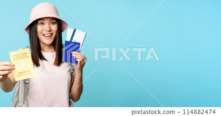 Enthusiastic asian tourist, girl shows covid-19 international vaccination certificate, passport with two tickets, travelling after coronavirus vaccine, blue background Enthusiastic asian tourist, girl shows covid-19 international vaccination certificate, passport with two tickets, travelling after coronavirus vaccine, blue background 114882474