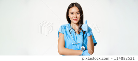 Smiling nurse, asian female doctor in scrubs, showing thumbs up sign and vaccinated arm with medical plaster, recommending vaccination from covid-19, white background 114882521