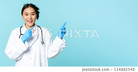 Happy smiling nurse, asian female doctor pointing right, showing promo, clinic advertisement, standing in medical uniform over blue background Happy smiling nurse, asian female doctor pointing right, showing promo, clinic advertisement, standing in medical uniform over blue background 114882548