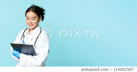 Asian female nurse or doctor writing down on clipboard with pen, smiling while examining patients, standing in uniform over blue background Asian female nurse or doctor writing down on clipboard with pen, smiling while examining patients, standing in uniform over blue background 114882567