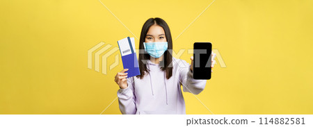 Happy korean girl in face mask, showing smartphone screen, app interface and passport with tickets for vacation, booking trip, yellow background Happy korean girl in face mask, showing smartphone screen, app interface and passport with tickets for vacation, booking trip, yellow background 114882581