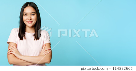 Smiling asian girl in t-shirt, holding arms crossed, ready for action pose, standing over blue background Smiling asian girl in t-shirt, holding arms crossed, ready for action pose, standing over blue background 114882665