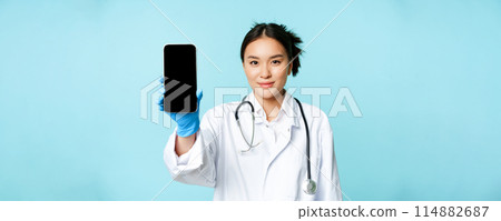 Web medical doctor concept. Smiling asian physician showing mobile phone screen, standing in clinic uniform, blue background Web medical doctor concept. Smiling asian physician showing mobile phone screen, standing in clinic uniform, blue background 114882687