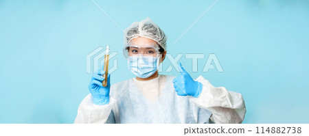 Asian female doctor, lab worker in personal protective equipment, showing thumbs up and test sample tube analise, standing over blue background Asian female doctor, lab worker in personal protective equipment, showing thumbs up and test sample tube analise, standing over blue background 114882738