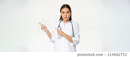 Sad and concerned female doctor, pointing fingers left, showing smth important, promo information, standing over white background Sad and concerned female doctor, pointing fingers left, showing smth important, promo information, standing over white background 114882753