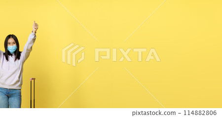 Full length shot of happy asian tourist, girl on vacation in medical face mask, showing thumbs up and standing near suitcase, yellow background 114882806