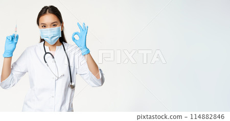Covid-19 vaccination and healthcare. Asian woman doctor in medical face mask and gloves, holding vaccine syringe and show okay sign, white background Covid-19 vaccination and healthcare. Asian woman doctor in medical face mask and gloves, holding vaccine syringe and show okay sign, white background 114882846