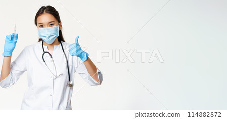 Covid-19 vaccination and healthcare. Asian woman doctor in medical face mask and gloves, holding vaccine syringe and show thumb up, white background 114882872