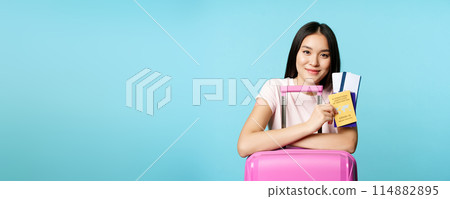 Happy asian girl with suitcare, shows coronavirus international vaccination certificate, two tickets for travelling abroad, going on trip after covid-19 vaccine, blue background 114882895
