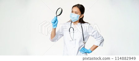 Image of asian female doctor, scientist in medical mask and rubber gloves looks through magnifying glass, searching for smth, white background Image of asian female doctor, scientist in medical mask and rubber gloves looks through magnifying glass, searching for smth, white background 114882949