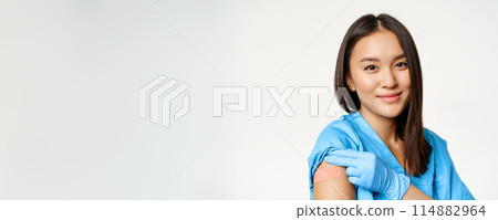 Vaccination program and covid-19 concept. Portrait of asian healthcare worker in medical robe, showing vaccinated arm and smiling, standing over white background Vaccination program and covid-19 concept. Portrait of asian healthcare worker in medical robe, showing vaccinated arm and smiling, standing over white background 114882964