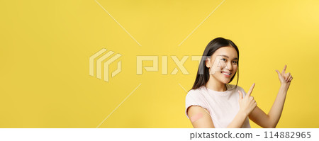 Covid-19 vaccination concept. Cute vaccinated asian woman demonstrating banner, pointing fingers at empty space for promotional text, yellow background 114882965