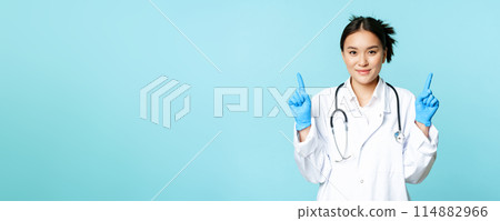 Smiling asian female doctor, nurse in medical uniform, gloves, pointing fingers up, showing healthcare information, standing over blue background Smiling asian female doctor, nurse in medical uniform, gloves, pointing fingers up, showing healthcare information, standing over blue background 114882966