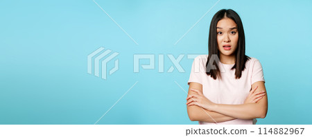 Portrait of young asian woman listening with confused, shocked face expression, standing in t-shirt over blue background Portrait of young asian woman listening with confused, shocked face expression, standing in t-shirt over blue background 114882967