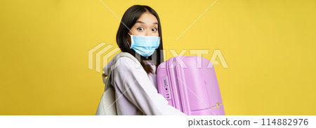Surprised asian girl tourist, wear medical face mask, looking amazed and impressed at camera, carry suitcase in hands, yellow background 114882976