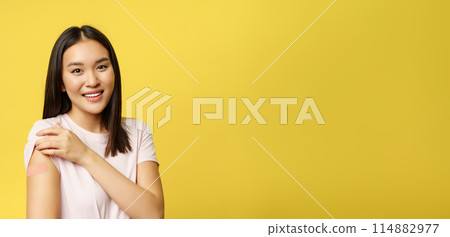 Covid-19 and healthcare medical concept. Smiling woman shows shoulder with patch, vaccinated from coronavirus, standing in t-shirt over yellow background 114882977