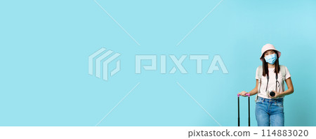 Smiling korean girl on vacation during pandemic. Standing with suitcase and photo camera, wearing medical face mask, blue background 114883020