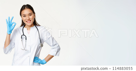 Smiling asian female physician, nurse in rubber gloves and medical uniform shows okay sign, approves and likes smth good, standing over white background 114883055