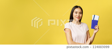 Smiling vaccinated from covid asian girl, showing passport with tickets, getting coronavirus vaccine shot for travelling abroad, yellow background 114883099