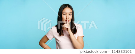 Smiling coquettish asian girl shushing, making silence taboo gesture, winking and making hush sign, press finger to lips, standing over blue background 114883101