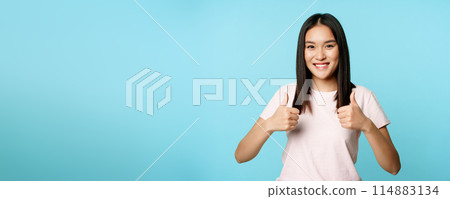 Enthusiastic asian girl showing thumbs up, like and approve, smiling satisfied, standing over blue background 114883134