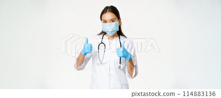 Covid-19 and medical workers concept. Smiling asian woman doctor in face mask and rubber gloves shows thumbs up, white background Covid-19 and medical workers concept. Smiling asian woman doctor in face mask and rubber gloves shows thumbs up, white background 114883136