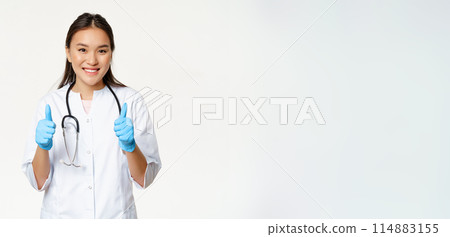 Smiling asian doctor, woman in medical uniform and rubber gloves shows thumbs up, recommending smth to patient, standing over white background Smiling asian doctor, woman in medical uniform and rubber gloves shows thumbs up, recommending smth to patient, standing over white background 114883155
