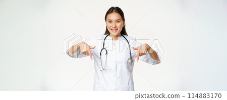 Smiling asian woman doctor, pointing fingers down and showing promo deal, discounts on clinic service, hospital advertisement, white background Smiling asian woman doctor, pointing fingers down and showing promo deal, discounts on clinic service, hospital advertisement, white background 114883170