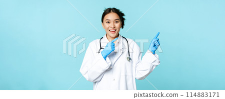 Happy smiling nurse, asian female doctor pointing right, showing promo, clinic advertisement, standing in medical uniform over blue background Happy smiling nurse, asian female doctor pointing right, showing promo, clinic advertisement, standing in medical uniform over blue background 114883171