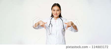 Young asian female doctor, pointing fingers down and smiling, wearing stethoscope and medical uniform, standing over white background Young asian female doctor, pointing fingers down and smiling, wearing stethoscope and medical uniform, standing over white background 114883172