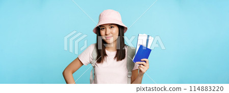 Smiling korean girl tourist, showing passport and tickets for flight, booking hotel, going on trip, voyage, standing over blue background Smiling korean girl tourist, showing passport and tickets for flight, booking hotel, going on trip, voyage, standing over blue background 114883220
