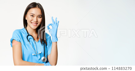 Smiling nurse, asian female doctor in scrubs, showing okay sign and vaccinated arm with medical plaster, recommending vaccination from covid-19, white background 114883270