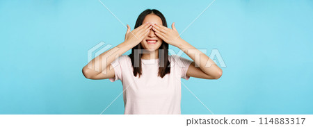 Happy smiling asian girl covers ears and waits for surprise, standing in t-shirt over blue background 114883317
