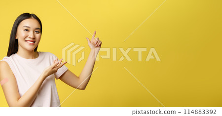 Covid-19 vaccination concept. Smiling asian vaccinated girl with patch on shoulder, pointing fingers at healthcare banner, yellow background 114883392