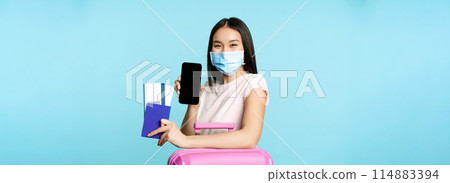 Tourism and healthcare concept. Asian female tourist with suitcare, wearing medical face mask for flight, showing smartphone screen and passport with tickets, vaccination certificate Tourism and healthcare concept. Asian female tourist with suitcare, wearing medical face mask for flight, showing smartphone screen and passport with tickets, vaccination certificate 114883394