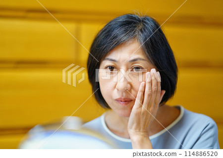 Middle-aged woman looking in the mirror and putting her hand on her cheek Middle-aged woman looking in the mirror and putting her hand on her cheek 114886520