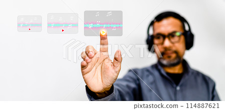 A man is wearing headphones and pointing at a screen with a music note on it 114887621