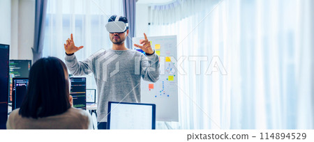Asian software developer wearing a virtual reality headset works on a VR project, with a colleague focused on her computer 114894529