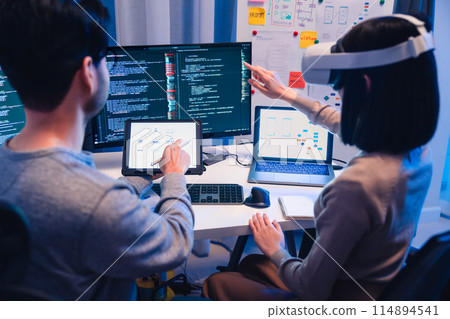 Asian software developer wearing a virtual reality headset works on a VR project, with a colleague focused on her computer Asian software developer wearing a virtual reality headset works on a VR project, with a colleague focused on her computer 114894541