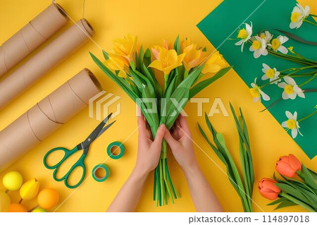 Florist making flower bouquet, hands holding flowers and scissors on table with colorful paper roll, yellow background, flat lay top view, copy space concept 114897018