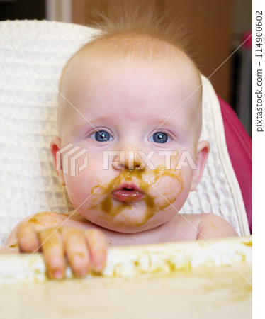 A cute baby with blue eyes has broccoli puree smeared all over their face. It looks like the baby is enjoying their meal while sitting at home. 114900602