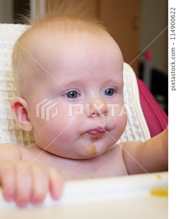 A cute baby with blue eyes has broccoli puree smeared all over their face. It looks like the baby is enjoying their meal while sitting at home. 114900622
