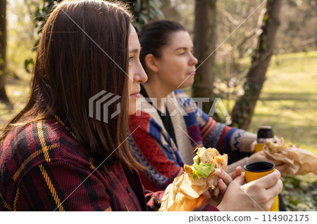 picnic in the woods, two young women eating and drinking tea from a thermos in the woods picnic in the woods, two young women eating and drinking tea from a thermos in the woods 114902175