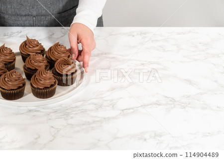 Freshly baked cupcakes have been masterfully infused with rich caramel and adorned with velvety chocolate frosting, all elegantly presented on a pristine white serving plate. 114904489