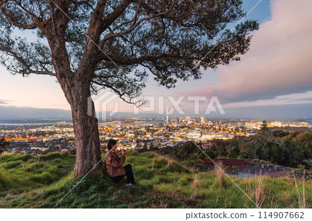 Viewpoint of Mount Eden with female tourist relaxing and sky tower among illuminated city at Auckland, New Zealand Viewpoint of Mount Eden with female tourist relaxing and sky tower among illuminated city at Auckland, New Zealand 114907662