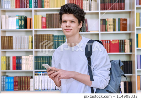 Teenage male student with backpack using smartphone, inside college building, in library 114908332