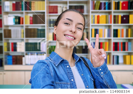 Selfie portrait of high school student girl showing hand gesture for victory, inside library 114908334