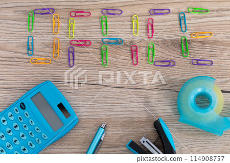 The inscription - office, made of colorful paper clips lying on a wooden desk top. Calculator and other office accessories. The inscription - office, made of colorful paper clips lying on a wooden desk top. Calculator and other office accessories. 114908757