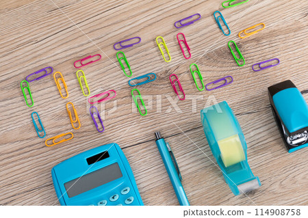 The inscription - office, made of colorful paper clips lying on a wooden desk top. Calculator and other office accessories. 114908758
