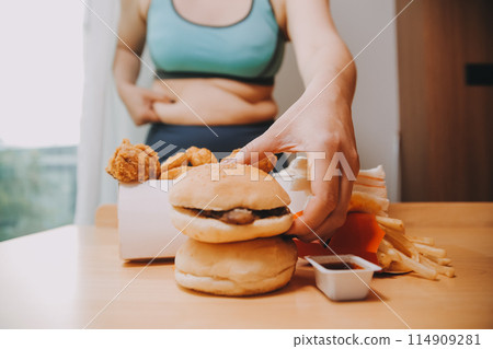 To eat or not. Cheerless plump woman looking at the burger while thinking to eat it or not To eat or not. Cheerless plump woman looking at the burger while thinking to eat it or not 114909281